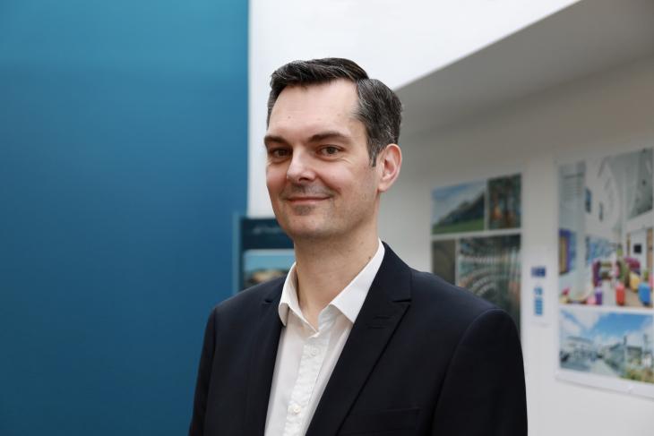 Robertson's Framework Manager Kevin Stewart, wearing a white shirt and dark suit, looking at the camera, in front of a blue wall.