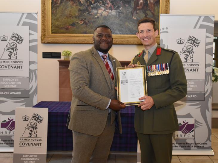 Robertson Group employee wearing a suit, collecting the Armed Forces Covenant Silver Award from a Brigadier with armed forces signage behind