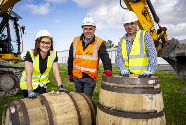 Robertson Construction Northern's MD Ian Phillips with Gordon & MacPhail employees in front of whisky casks with an excavator in the background