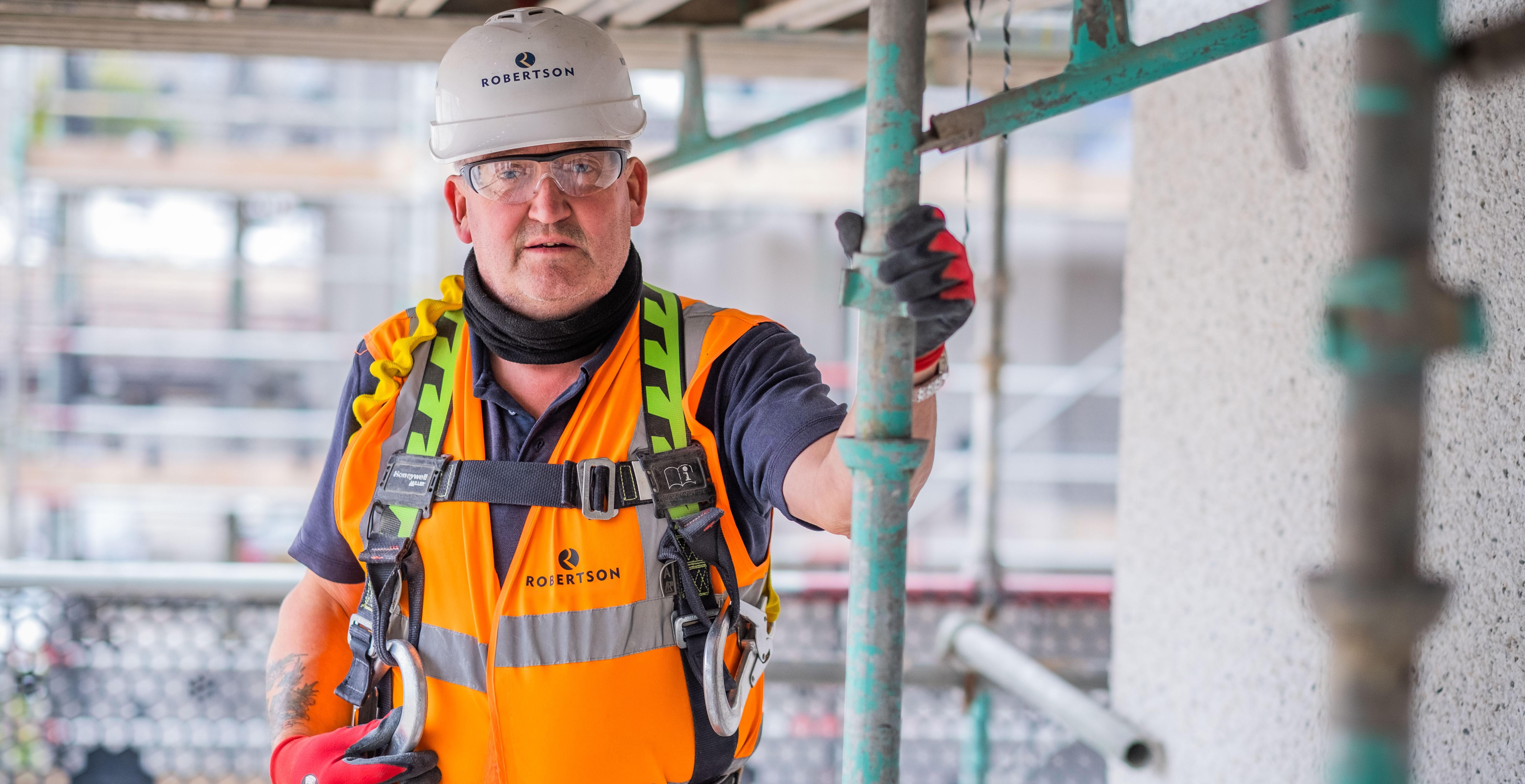 SHE image: Construction worker in full PPE including hard hat, protective goggles, orange high viz vest, gloves and safety harness