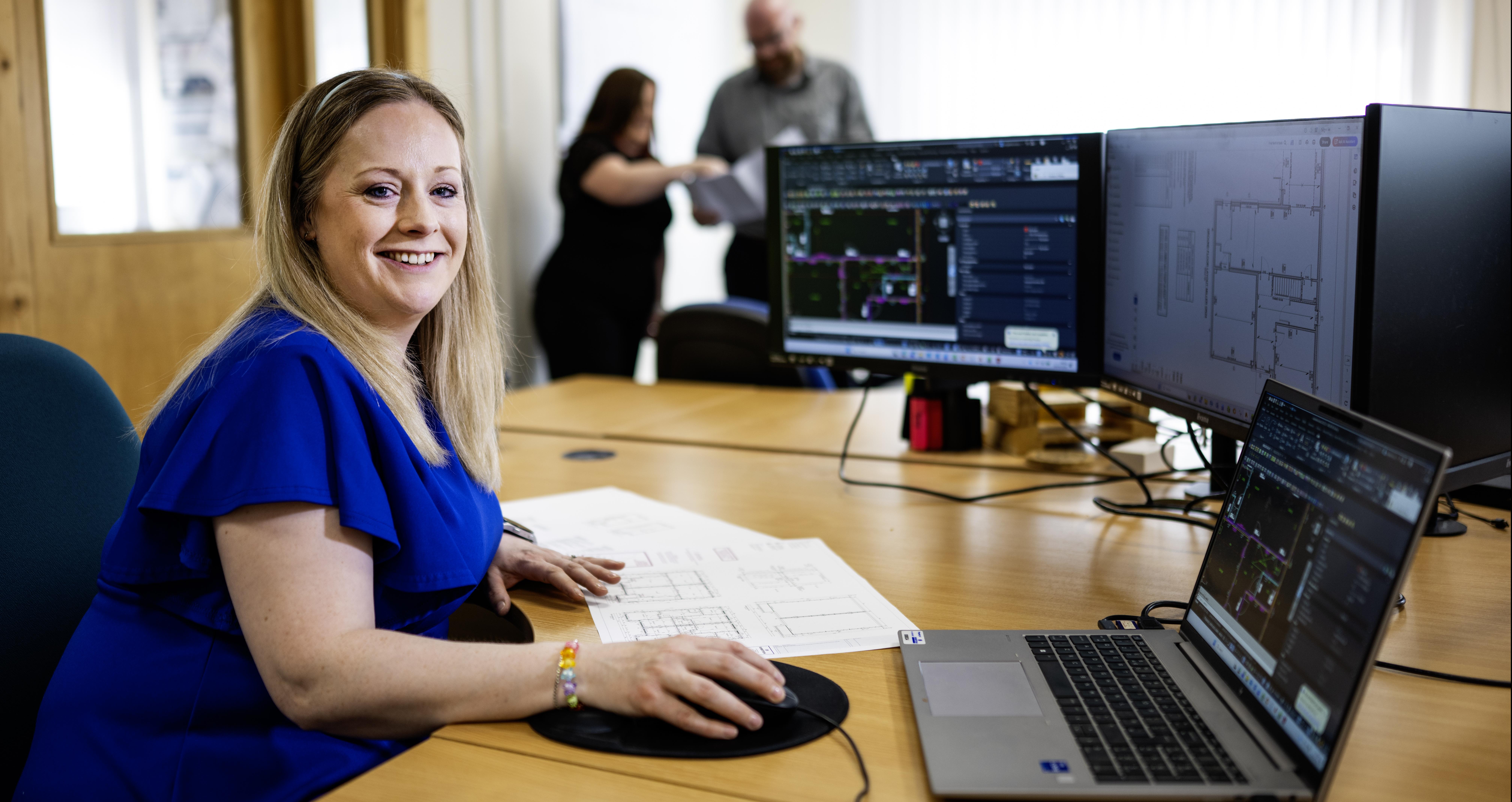 A woman smiling wearing blue dress sitting at a desk with computer design software