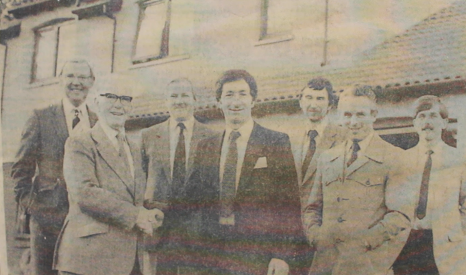An old black and white photograph of Bill Robertson in the early years of his company. He is pictured shaking hands with another man, surrounded by others who are all smiling at the camera.