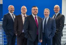 Five men in a mix of grey and navy suits stood together in front of a stairwell and blue wall, who are the new Robertson Partnership Homes Board 