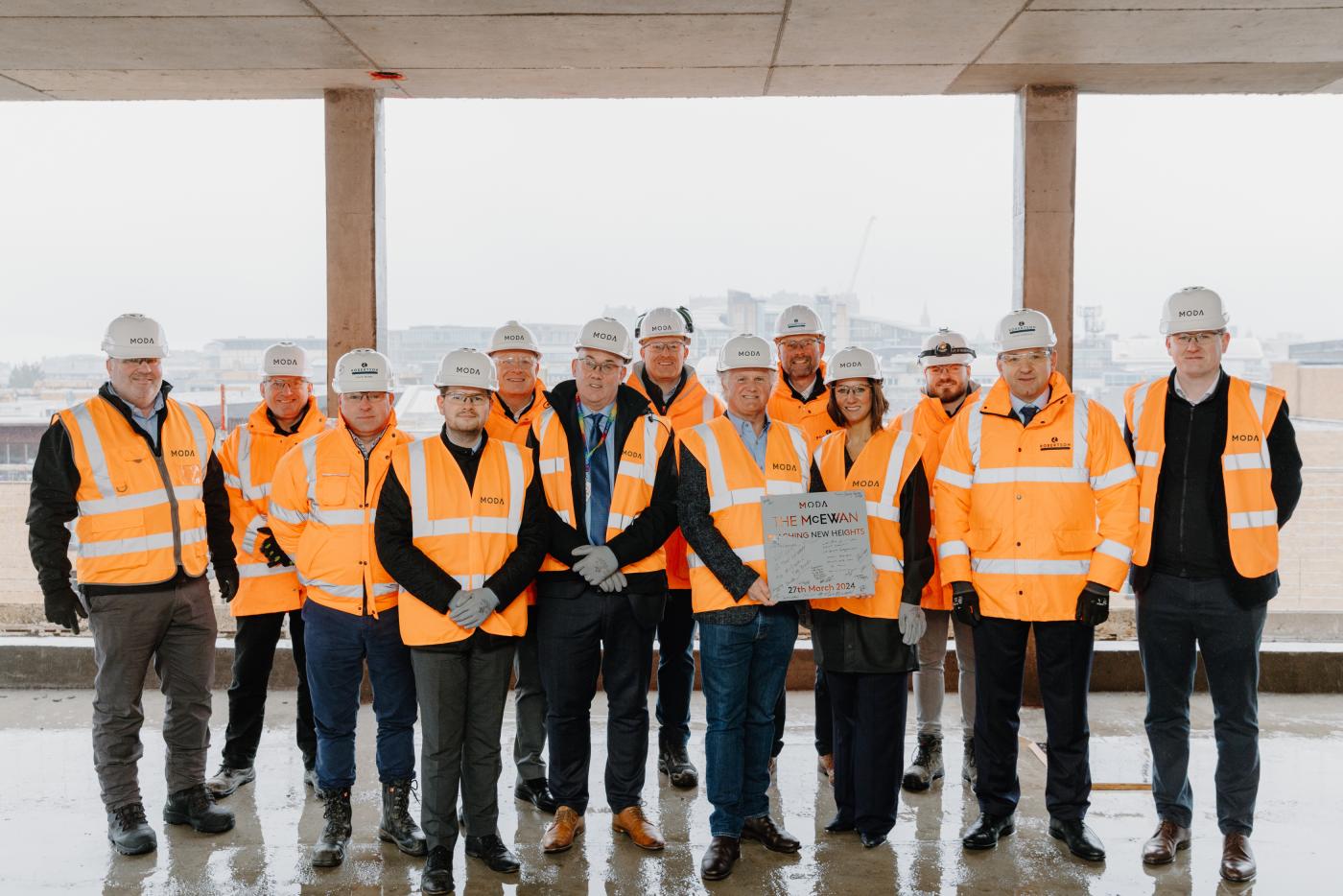 People gathered around two people holding a signed metal plaque to celebrate a construction topping out