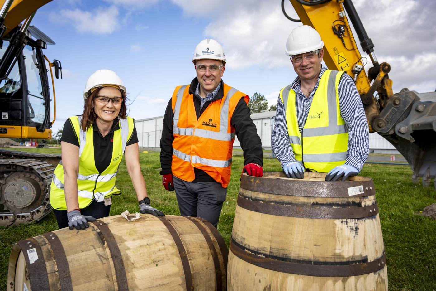 Robertson Construction Northern's MD Ian Phillips with Gordon & MacPhail employees in front of whisky casks with an excavator in the background