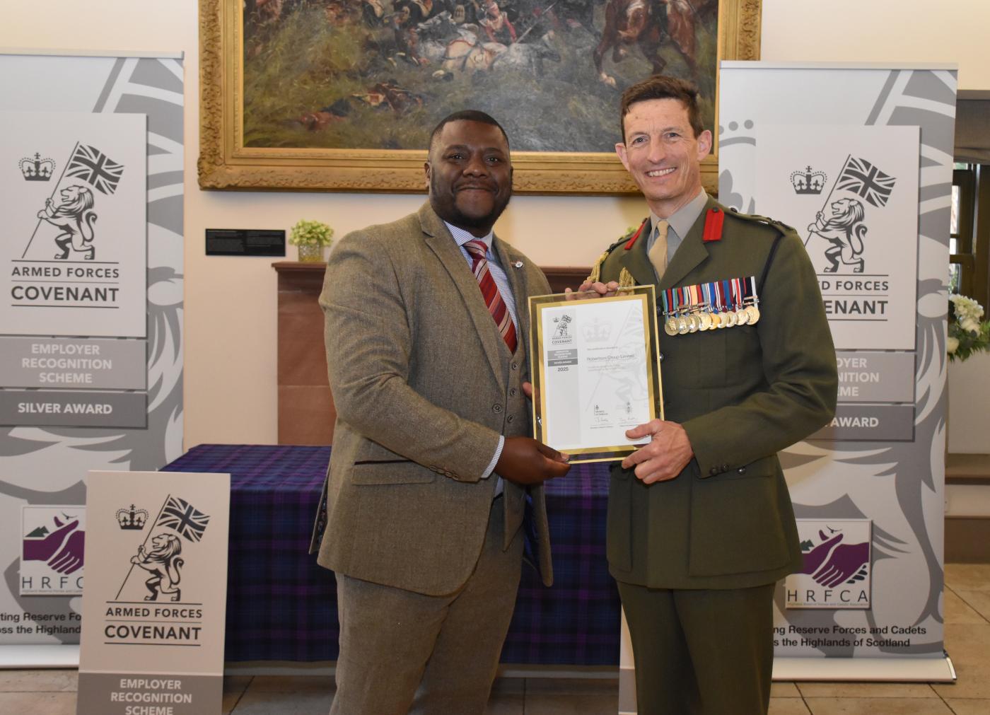 Robertson Group employee wearing a suit, collecting the Armed Forces Covenant Silver Award from a Brigadier with armed forces signage behind