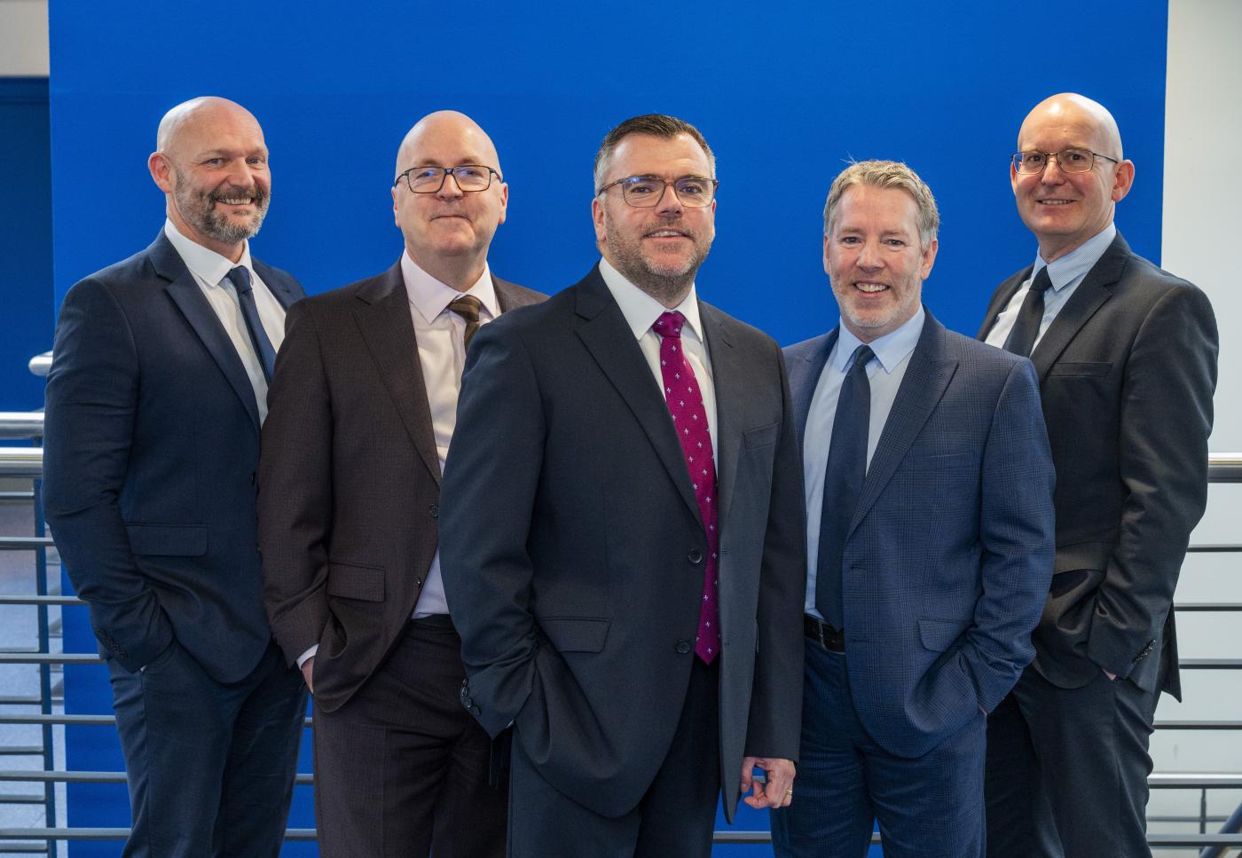Five men in a mix of grey and navy suits stood together in front of a stairwell and blue wall, who are the new Robertson Partnership Homes Board 